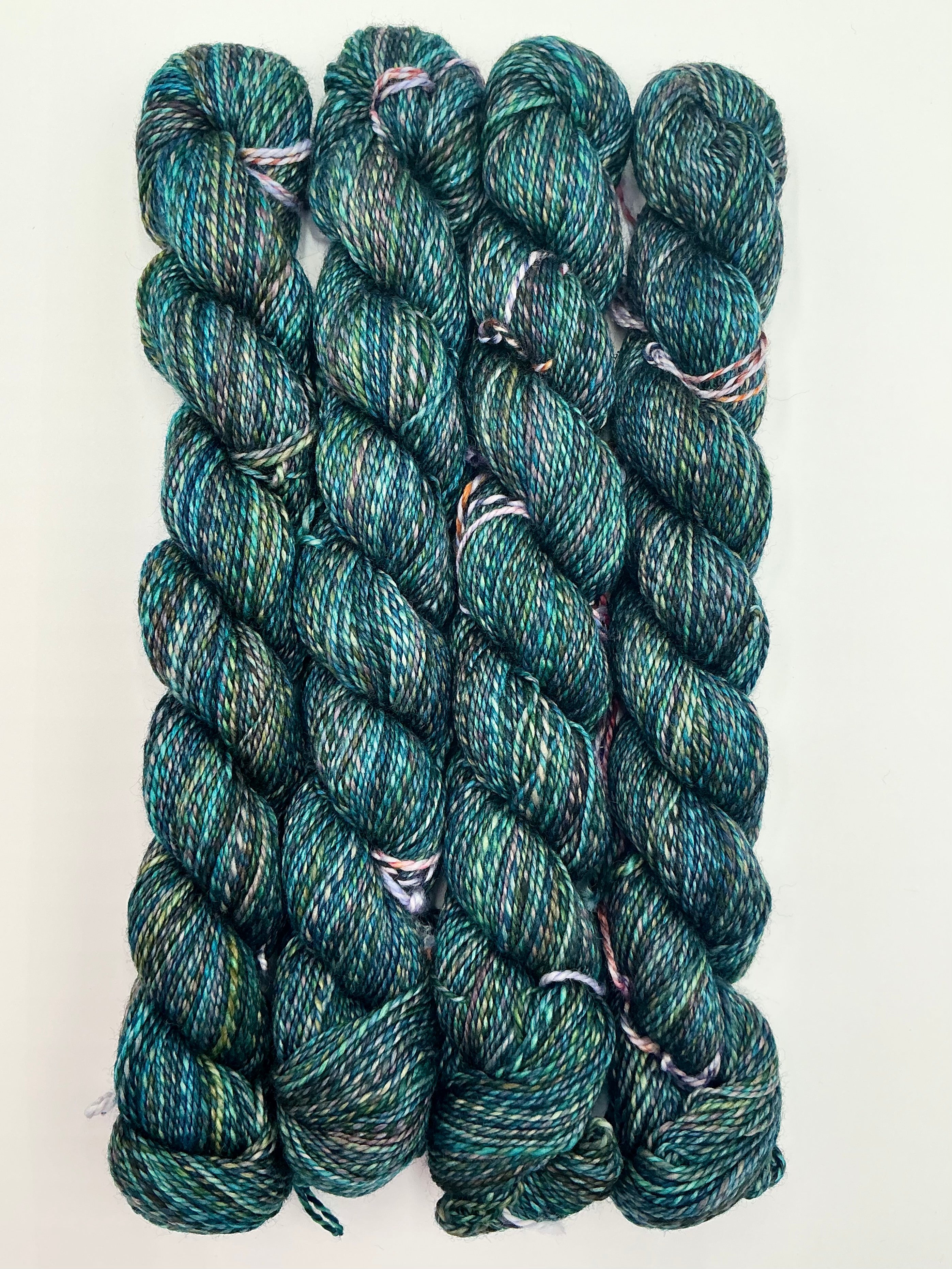 Four skeins of variegated yarn in a heathered teal, green, and blue colorway, arranged vertically on a plain white background.