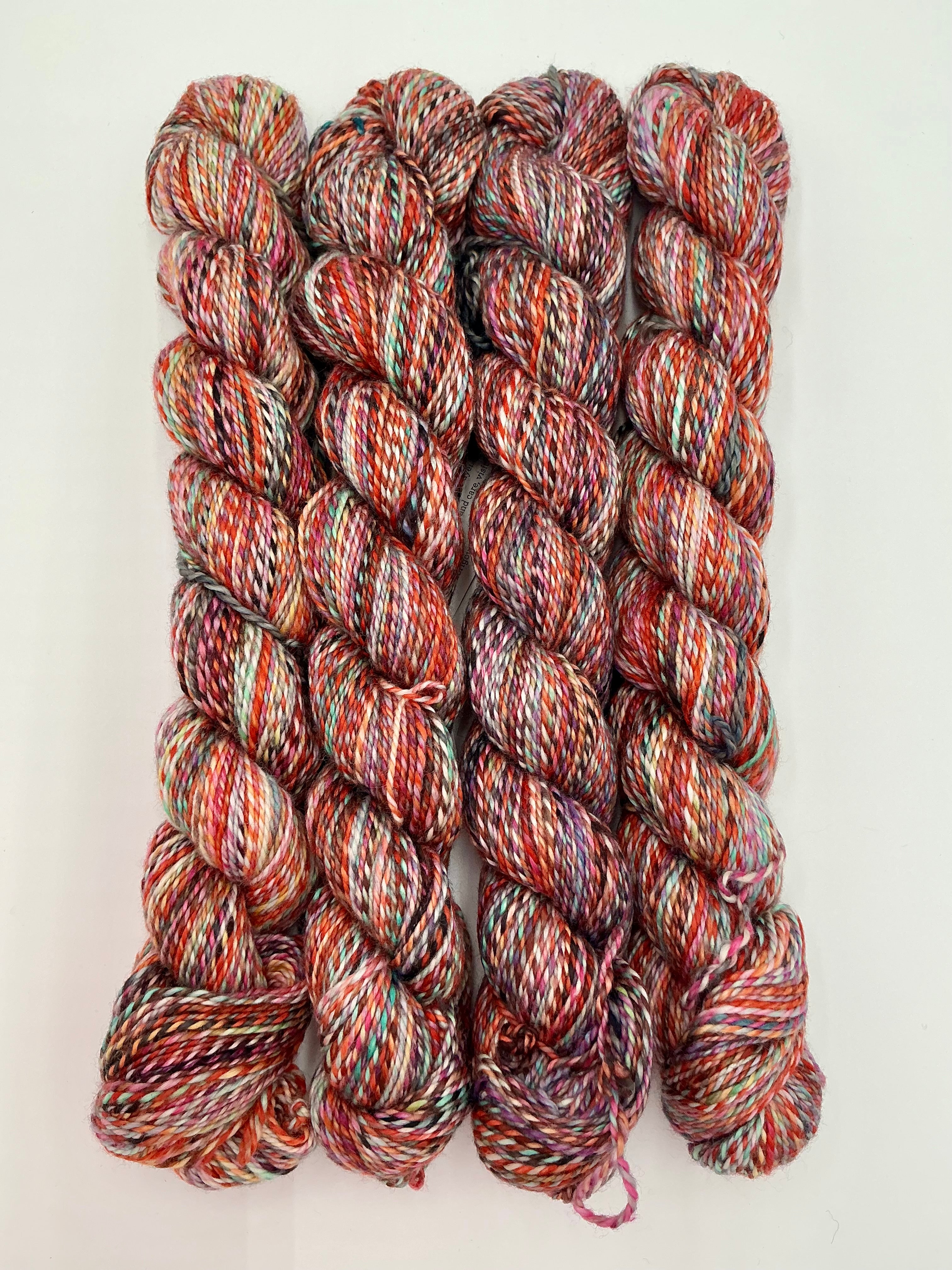 Four twisted skeins of variegated yarn, with a multicolor blend of red, orange, pink, green, and cream tones, arranged on a white background.