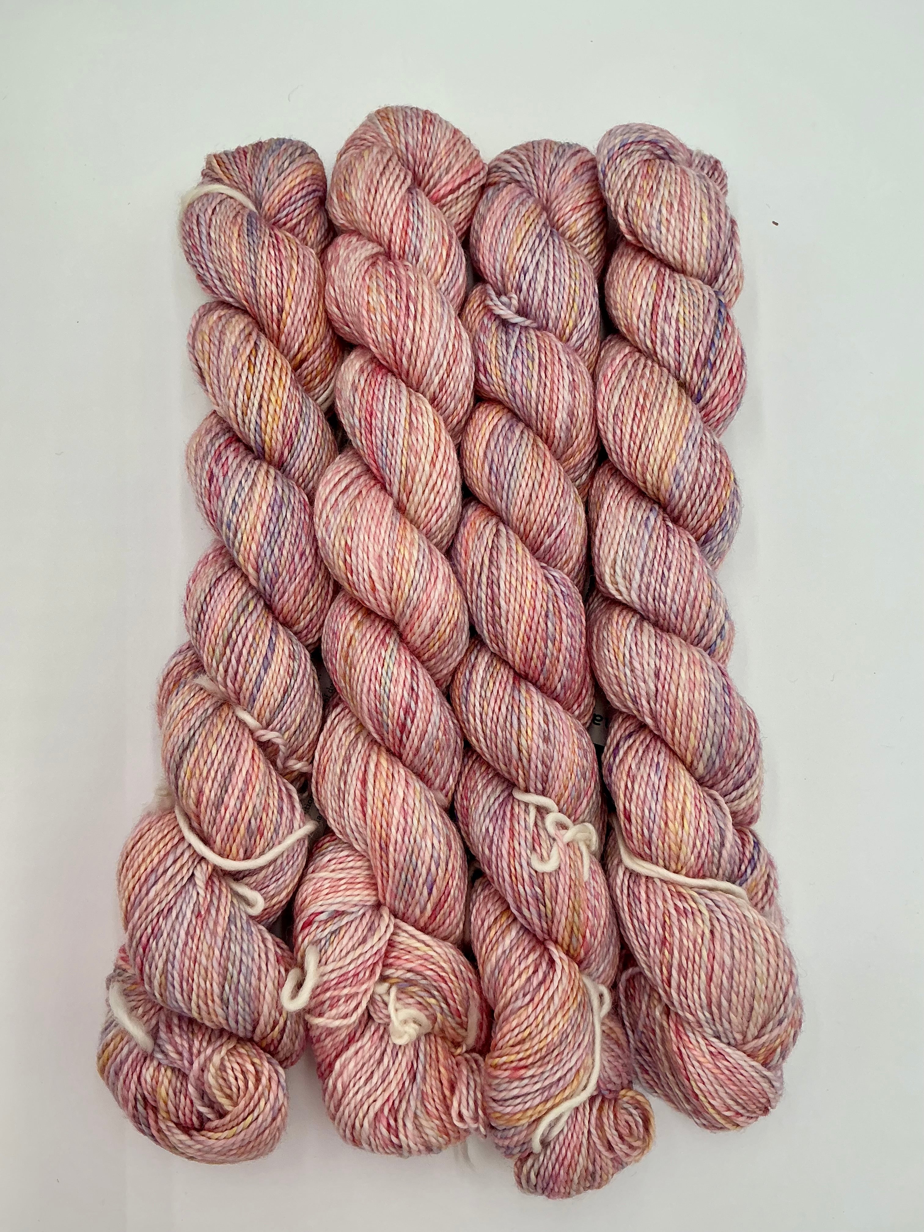 Four skeins of variegated yarn in dusty pink, mauve, and cream tones, arranged vertically on a white background.
