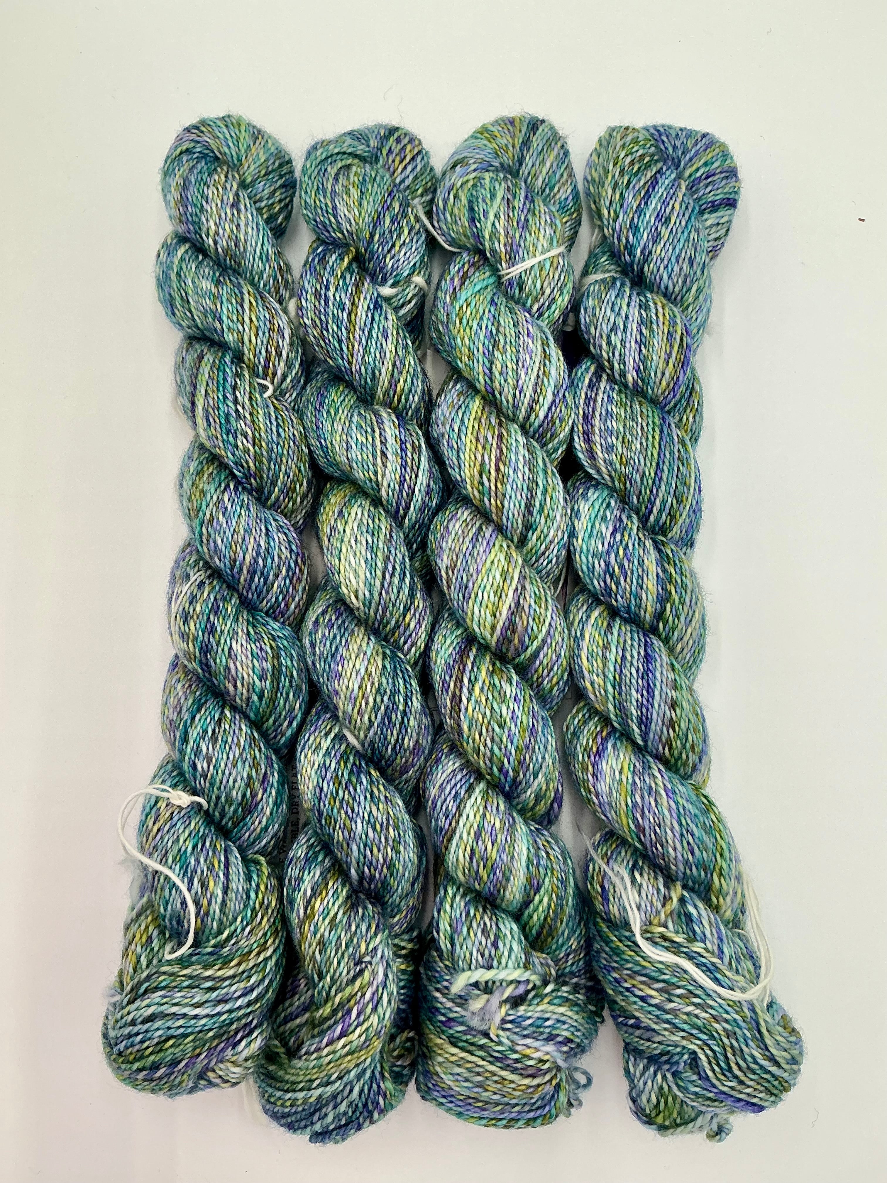 Four skeins of variegated yarn with a blend of blue, green, and purple tones, arranged vertically on a white background.