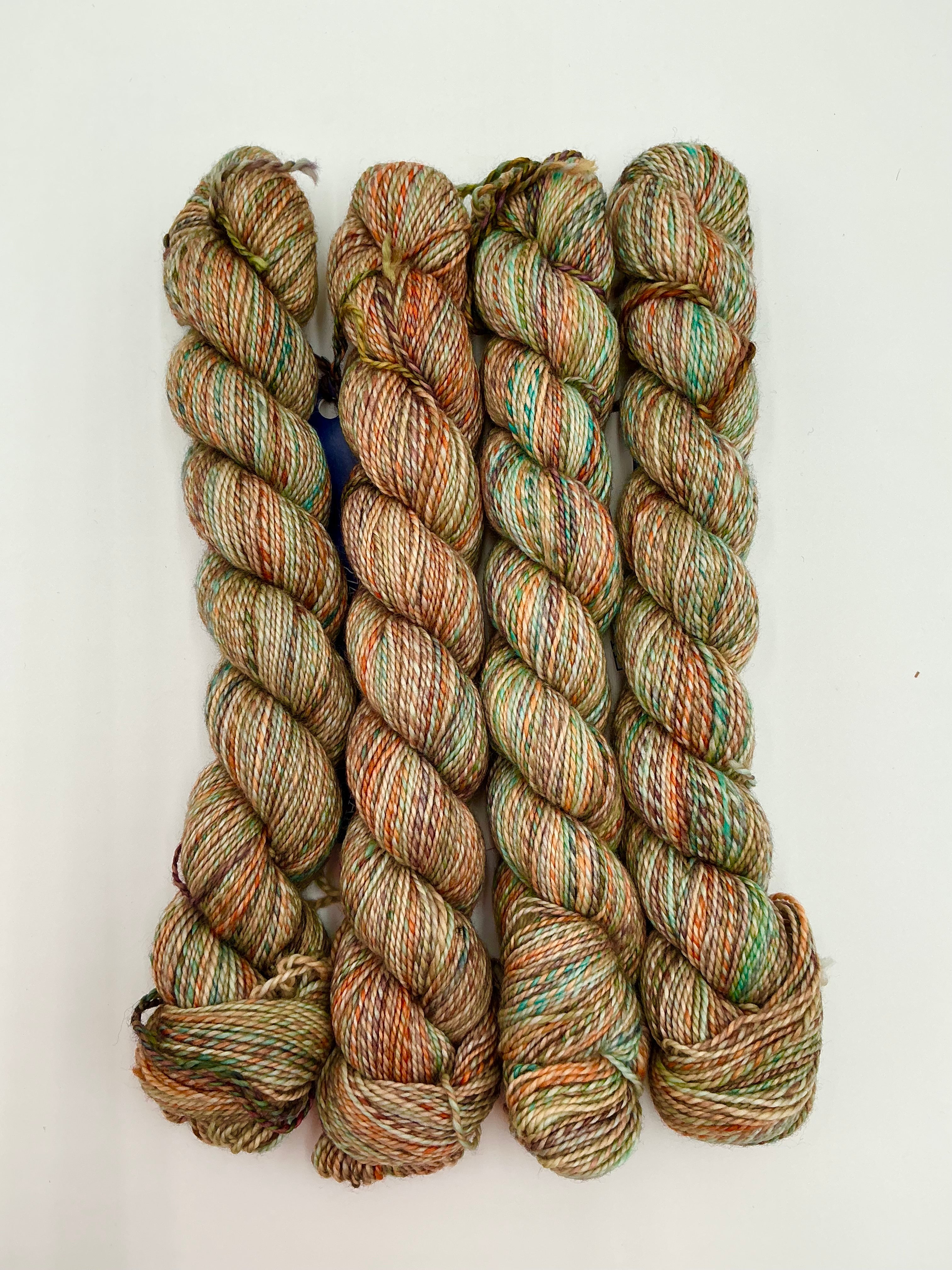 Four skeins of variegated yarn in earthy shades of beige, brown, muted green, and orange, arranged vertically on a plain white background.