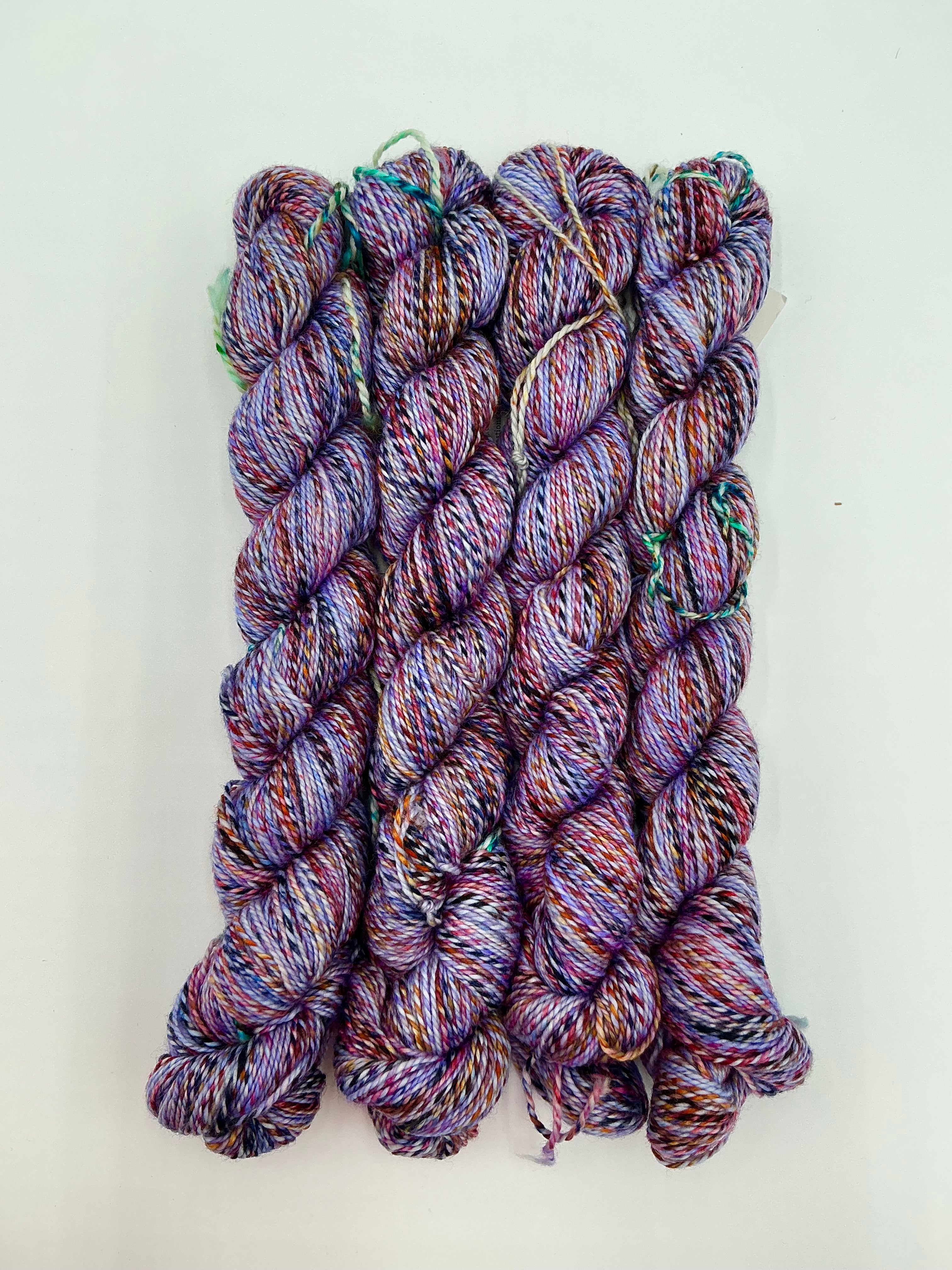 Four skeins of variegated yarn in shades of purple, pink, red, and black, arranged vertically on a plain white background.